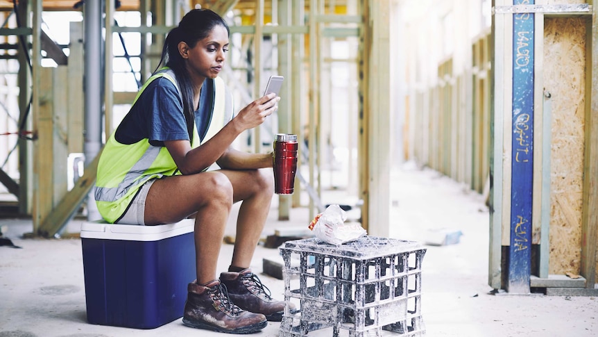 A young woman of colour sits looking at her phone during a work break on a construction site