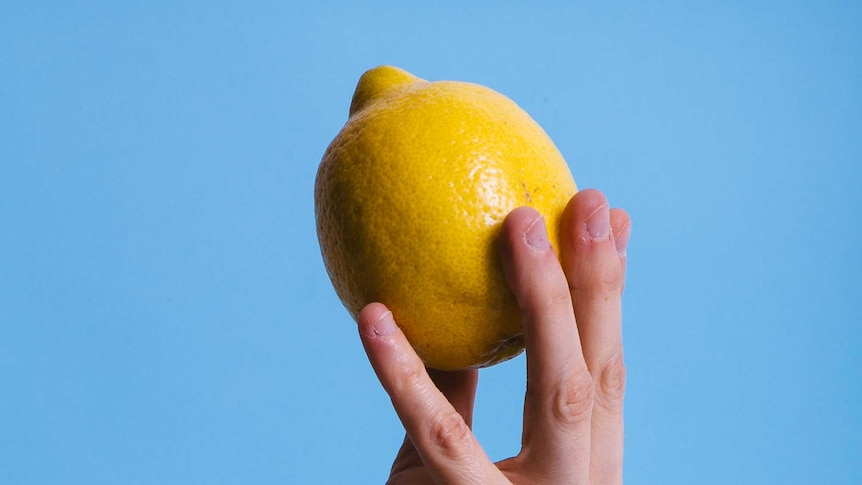Hand holding a bright yellow lemon up in the air against a pale blue background in a story about ways to use up lemons.