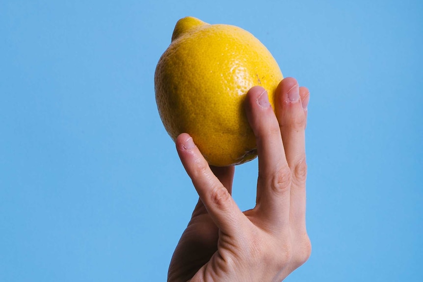 Hand holding a bright yellow lemon up in the air against a pale blue background in a story about ways to use up lemons.