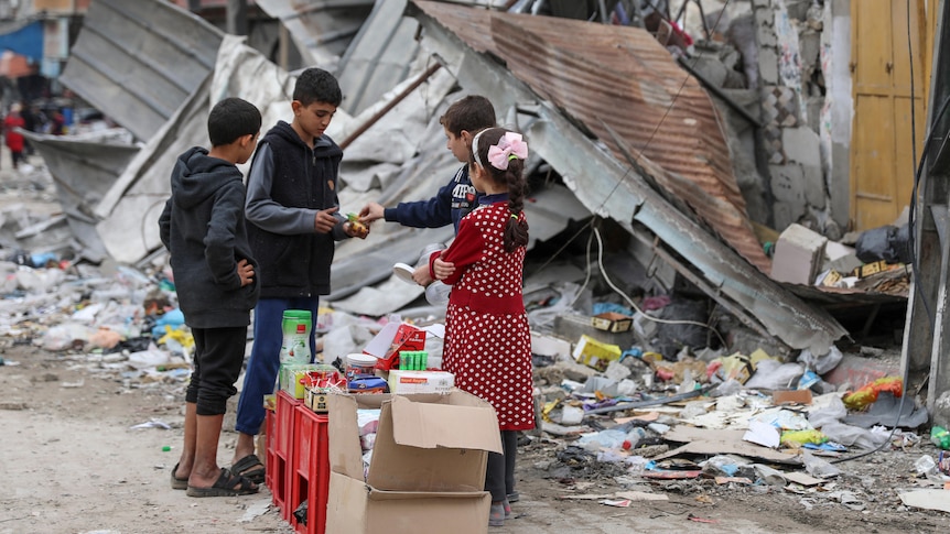 A wide shot of four children exchanging money and sweets on a street, in front of rubble and a badly damaged building.