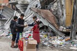 A wide shot of four children exchanging money and sweets on a street, in front of rubble and a badly damaged building.