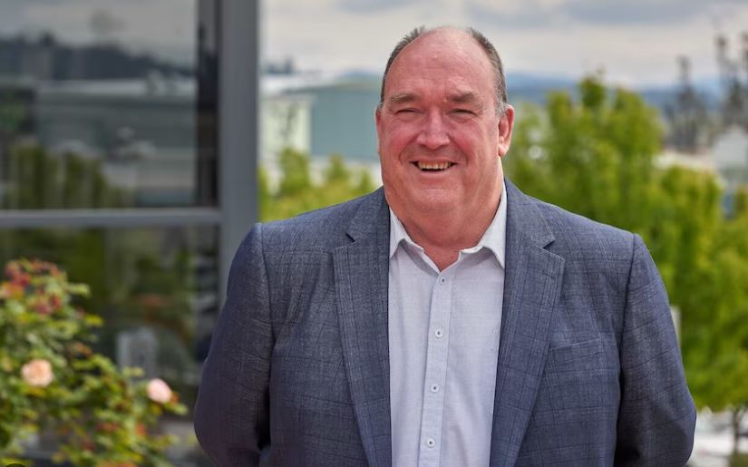A smiling man in a suit standing outside with trees and buildings behind him.