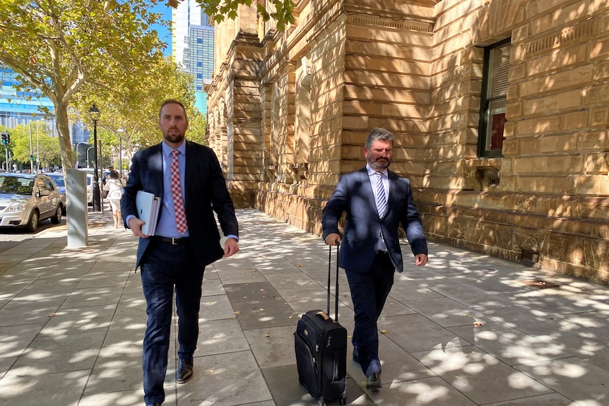 Two men in suits walk along a footpath, one pushing a small suitcase on wheels
