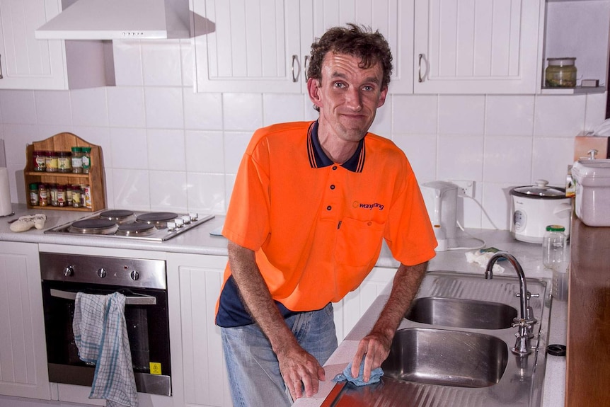 A man in a bright orange shirt uses a dishcloth to wipe a kitchen sink