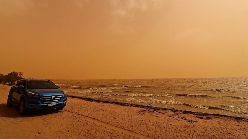 A blue car parked in front of a choppy lake, against the backdrop of a red sky due to a dust storm.