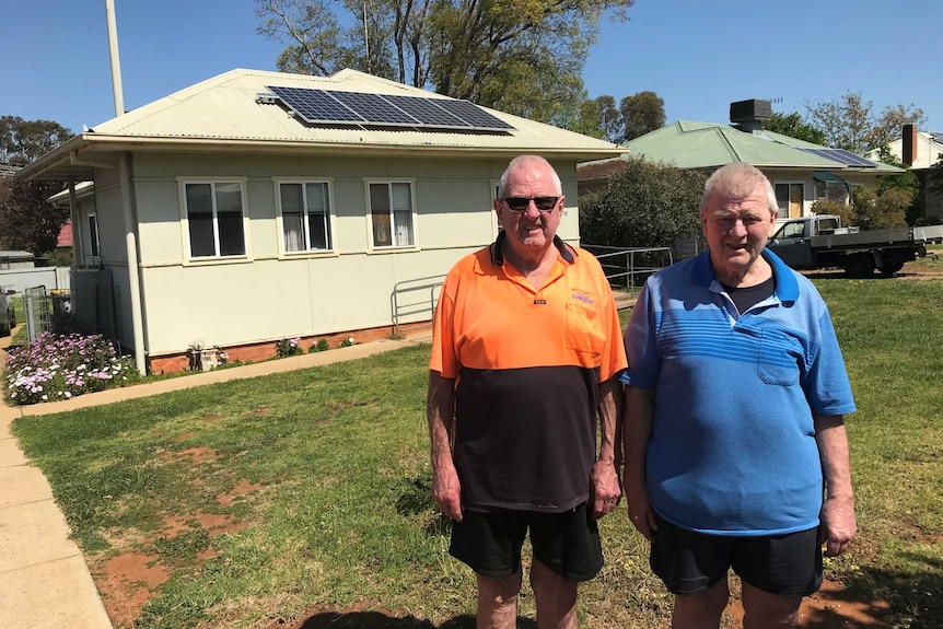 Two older men standing in front of a white house with a solar panel on the roof.