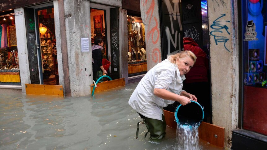 A woman removes water from a shop using a bucket in a flooded street of Venice.