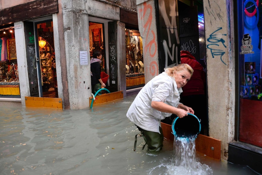 A woman removes water from a shop using a bucket in a flooded street of Venice.