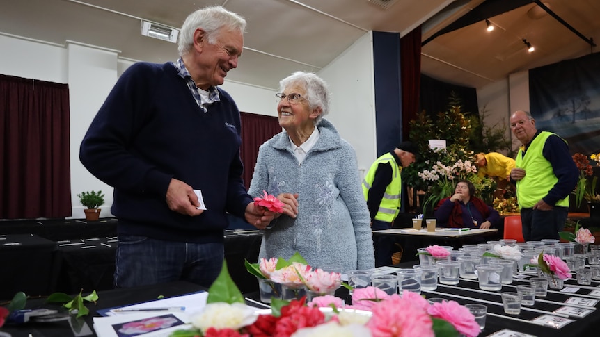 A man and a woman talking to each other in a hall. The man is holding the cut flower from a camellia