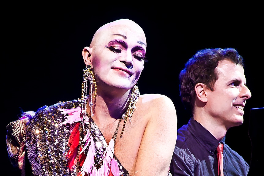 Taylor Mac, a bald white drag performer, and Matt Ray, a white queer man, sit at a piano and perform onstage.