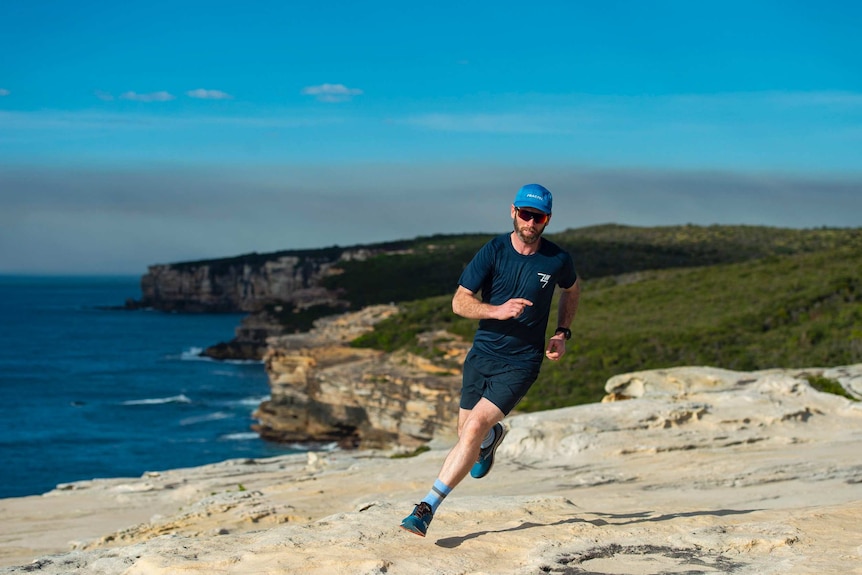 Man in shorts, t-shirt, cap and sunglasses running on cliff top overlooking beach