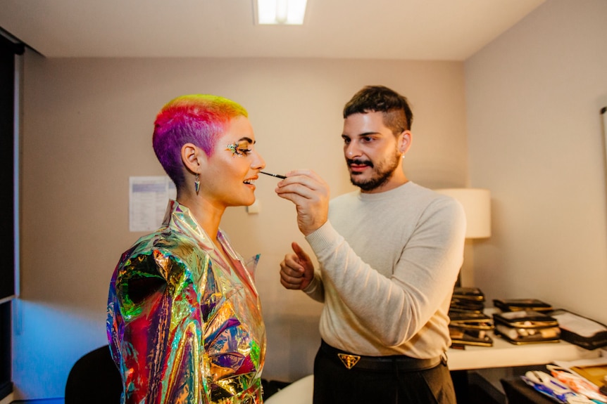A woman stands in an iridescent coat as another person in a beige jumper touches up her makeup. Her hair is multicolour.