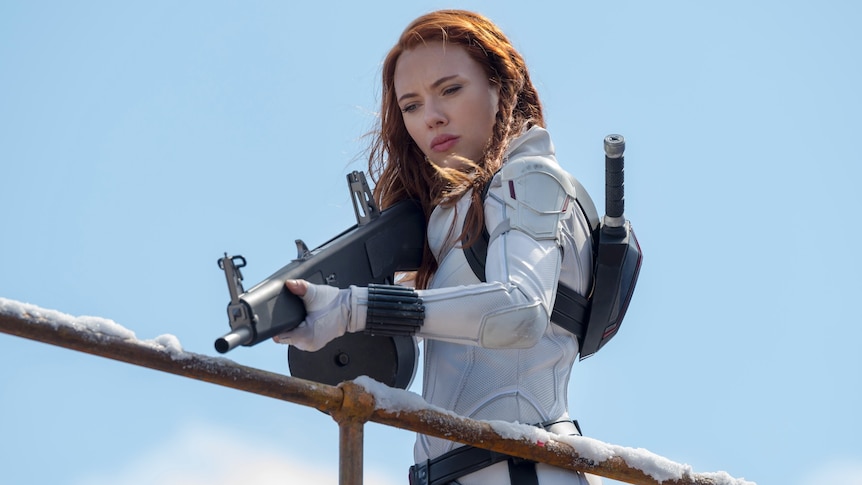 Scarlett Johansson in a white superhero suit points a gun downwards, over a fence