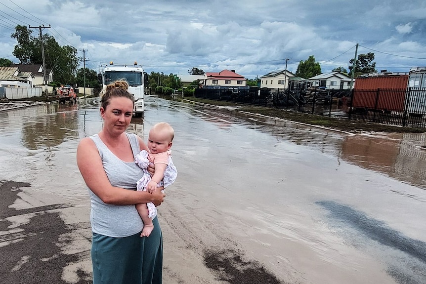 Mother stands in flooded suburban street holding baby.