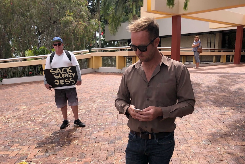 Jess Glasgow outside council's offices. A protester stands behind him with a sign that says "Sack sleazy Jess".