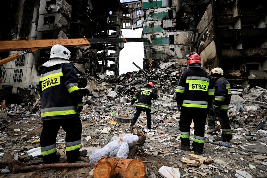 People in hard hats and uniforms search rubble from destroyed buildings.
