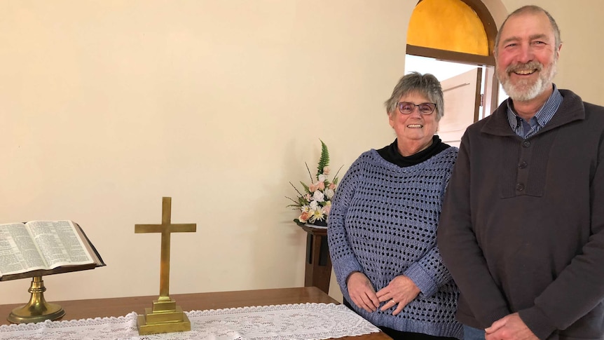 Smiling man and woman stand behind wooden table with golden cross and bible.