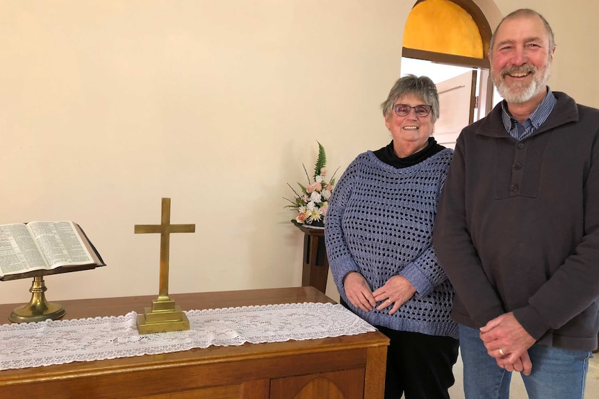 Smiling man and woman stand behind wooden table with golden cross and bible.