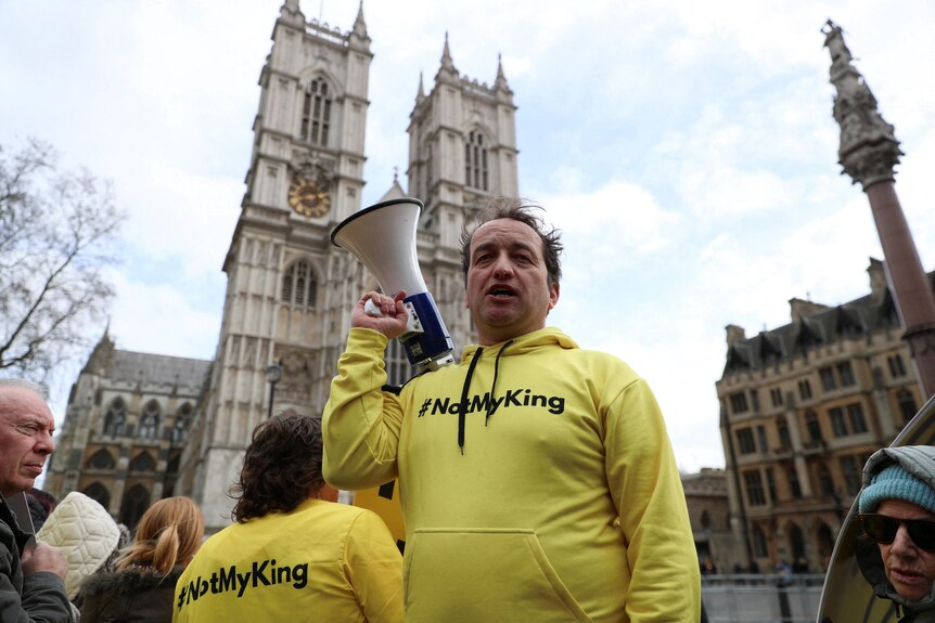 A man in a yellow hoodie with the words #NotMyKing stands with a megaphone in front of Westminster Abbey