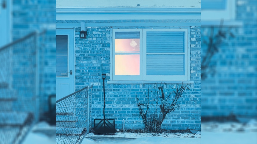 An icy blue photo of a brick house with a white-pink glow through the window, a snow shovel lies against the wall