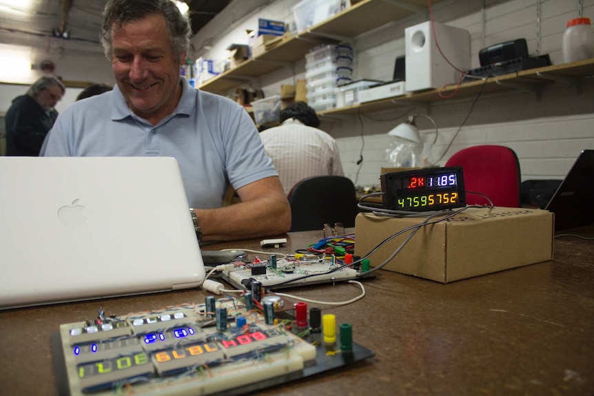 A man sits behind a laptop surrounded by alpha-numberic LED displays