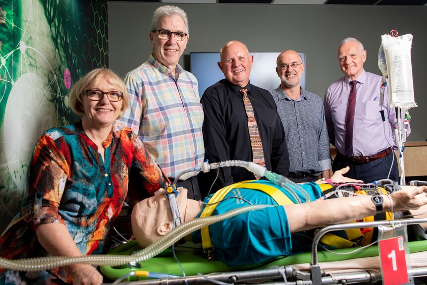 A group of people smiling while standing around a medical mannequin lying down.