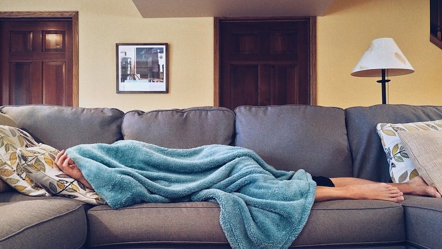 A person lies on a grey couch with a blue blanket pulled over their head and body. Feet are exposed.
