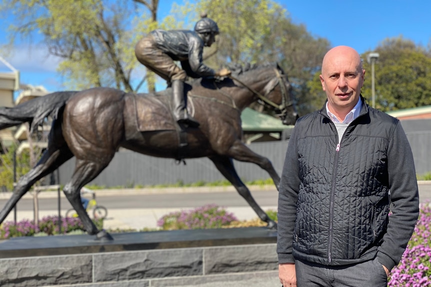 A bald man in front of a statue of a jockey and horse