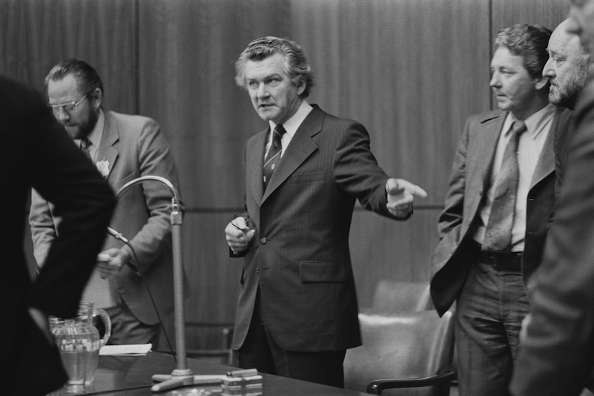 Bob hawke stands next to a table, pointing to his left, surrounded by other men.