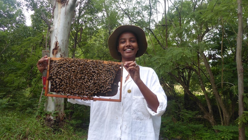 A woman wearing an oversized white shirt holds up an apparatus holding thousands of bees.