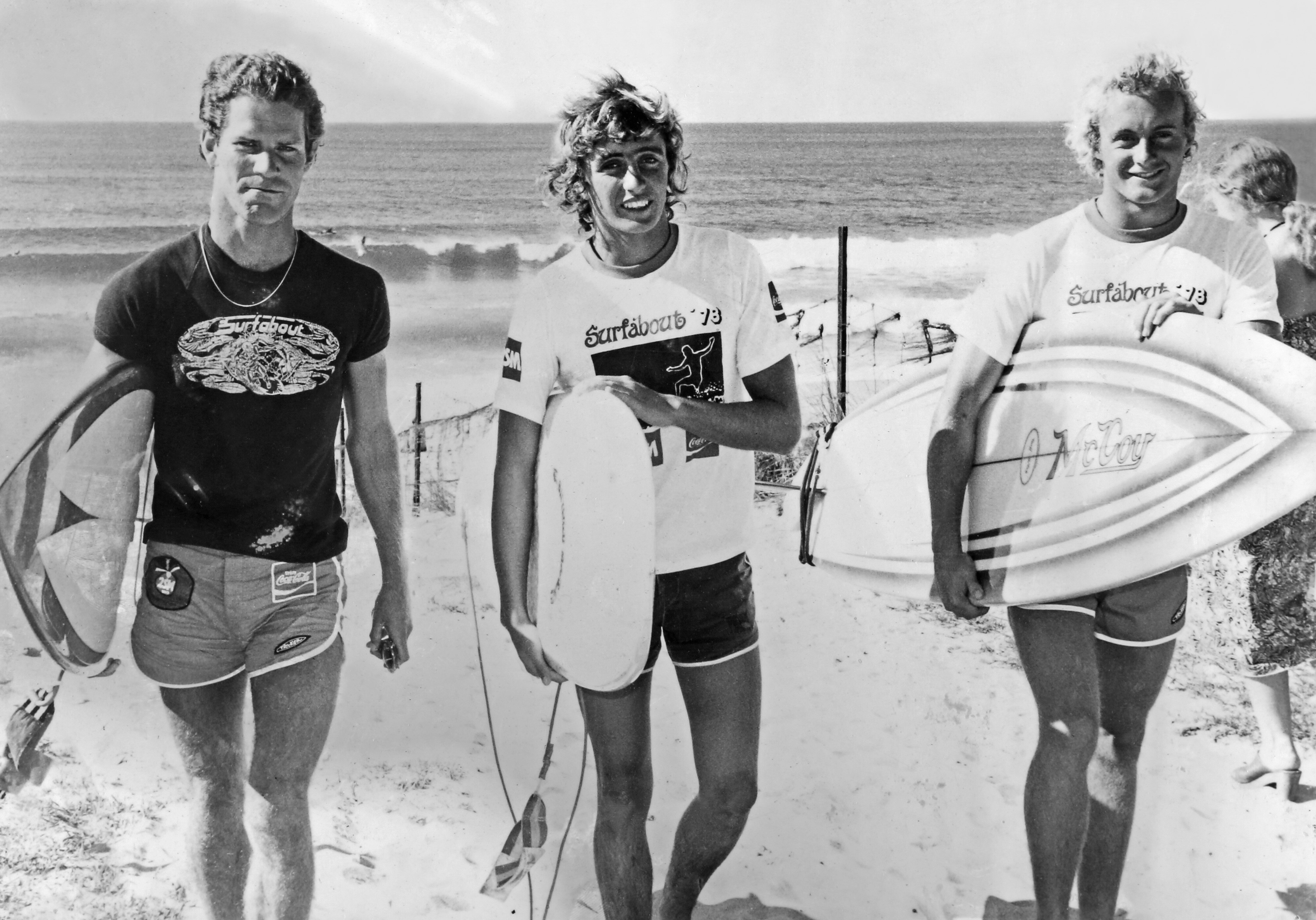 Black and white photo of three men in shorts and tee holding surf boards, smiling, ocean at the back.
