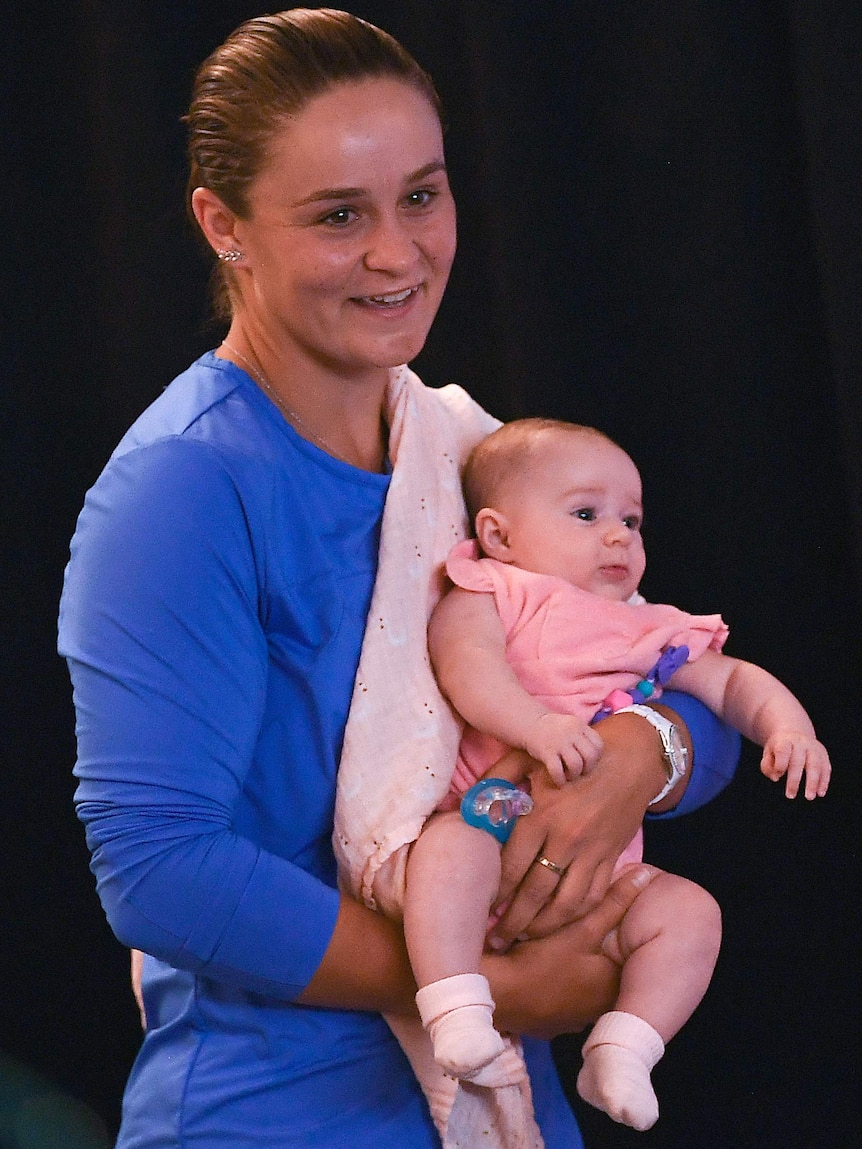 A smiling tennis player stands while holding a baby at the end of a press conference.