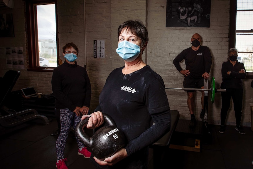 A woman in black clothes with a blue covid facemask holds a gym kettlebell