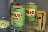 Three old green tins with retro font spelling Milo sit on shelf.