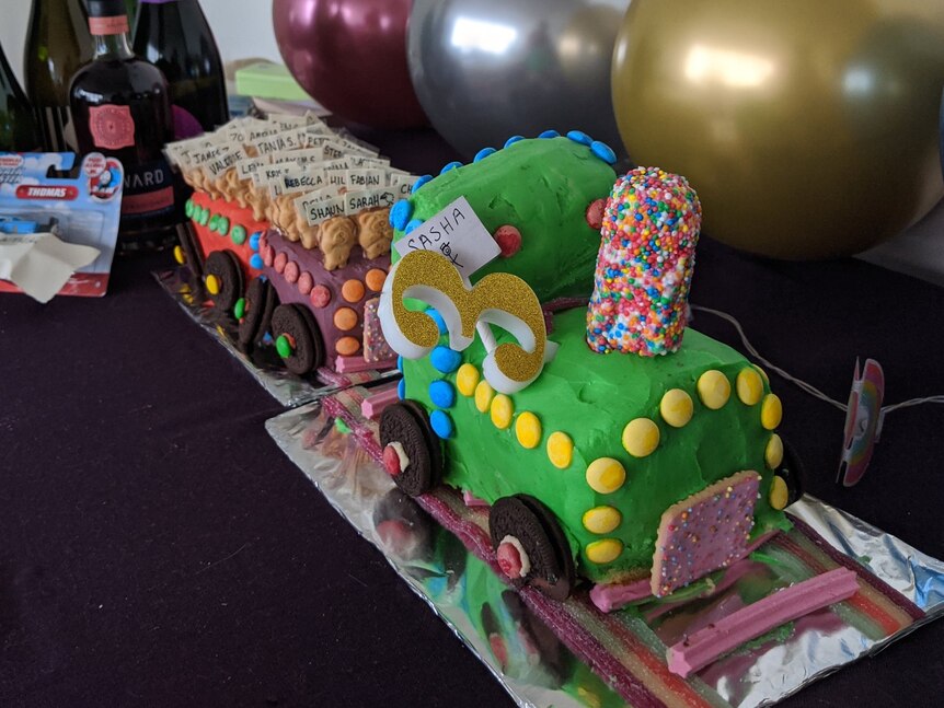 An elaborate Woman's Weekly birthday cake shaped like a train, on a table with balloons and other gifts.
