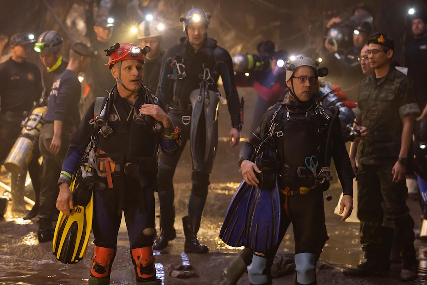 Three white men in scuba diving gear and torches stand in a dark cave surrounded by other divers.