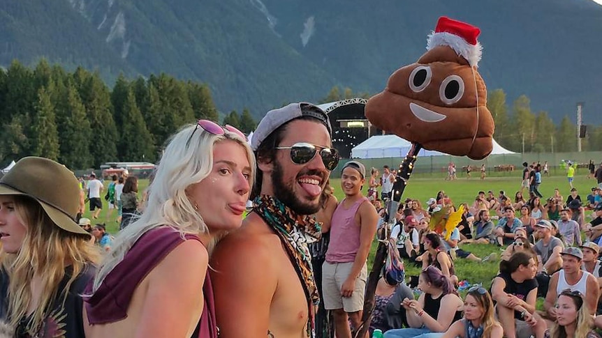 Two people at a music festival smile as they take a photo using a selfie stick with the smiling poop emoji attached.
