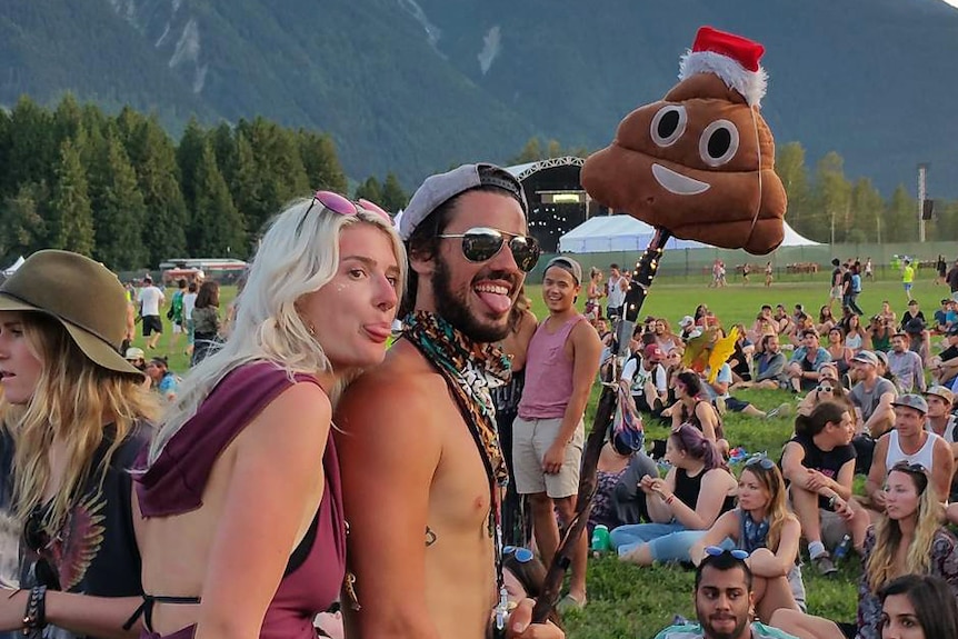 Two people at a music festival smile as they take a photo using a selfie stick with the smiling poop emoji attached.