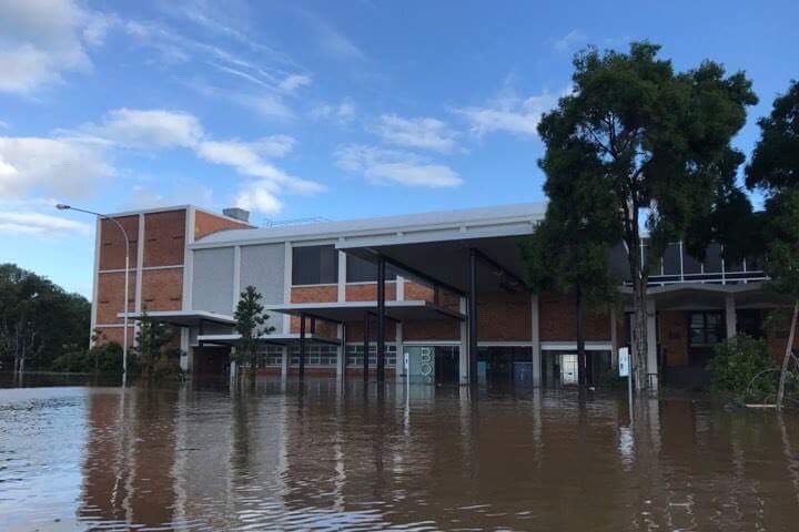 A large building, made of red brick, a multipurpose arts space, is surrounded by floodwater on a clear day