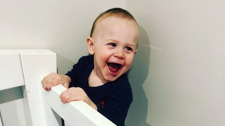 A smiling two-year-old boy holds onto the side of his cot.