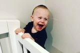 A smiling two-year-old boy holds onto the side of his cot.