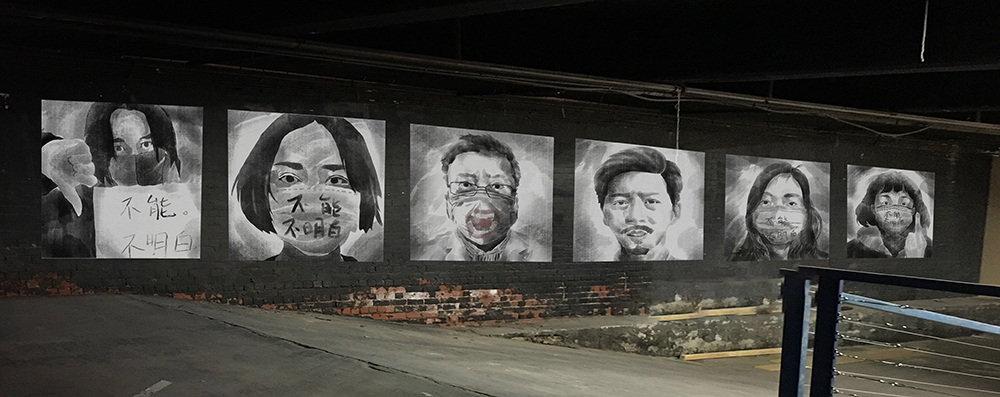 Six black and white water colour style portraits of people wearing face masks are hung on black warehouse wall.