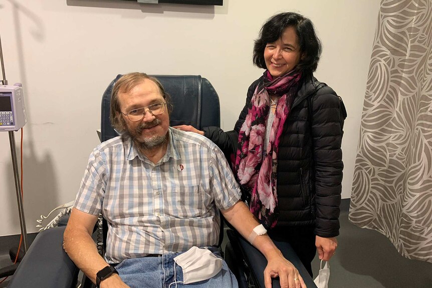 A man sits in a chair and a woman stands with her hand on his shoulder.