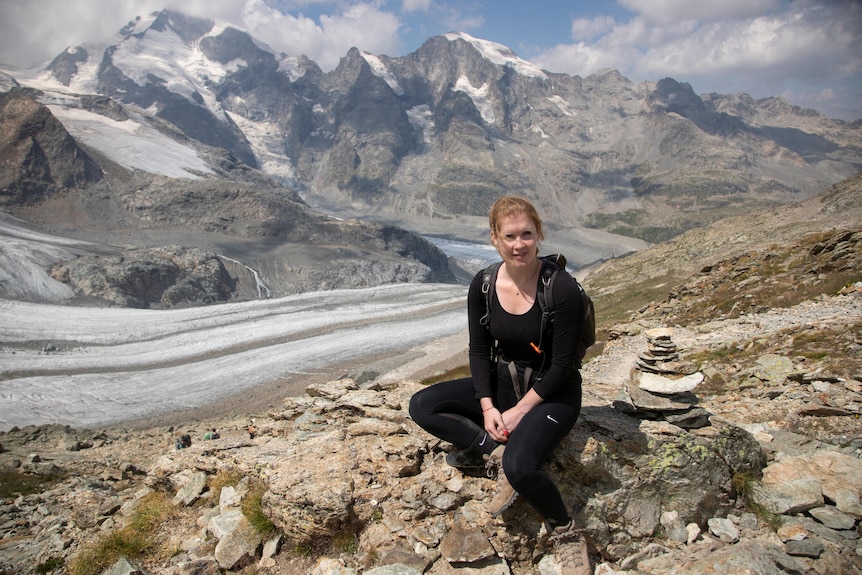 A woman is crouched down at smiling at the camera with the Swiss alps in the background. 