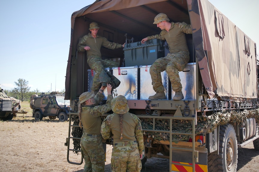 Two soldiers sit in the back of an army truck while two others pass them equipment