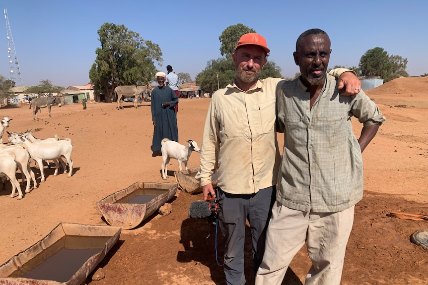 Cameraman with arm around another man and goats in background.