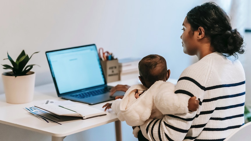 Woman with a striped jumper works on a laptop while holding a baby at a desk in her home.