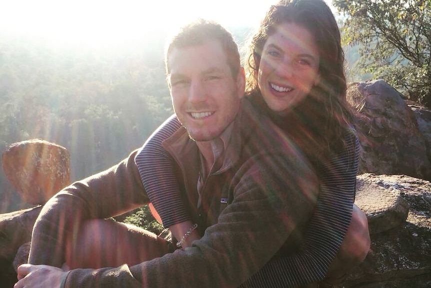 A man and woman smile while hugging. The sunlight beams behinhd them