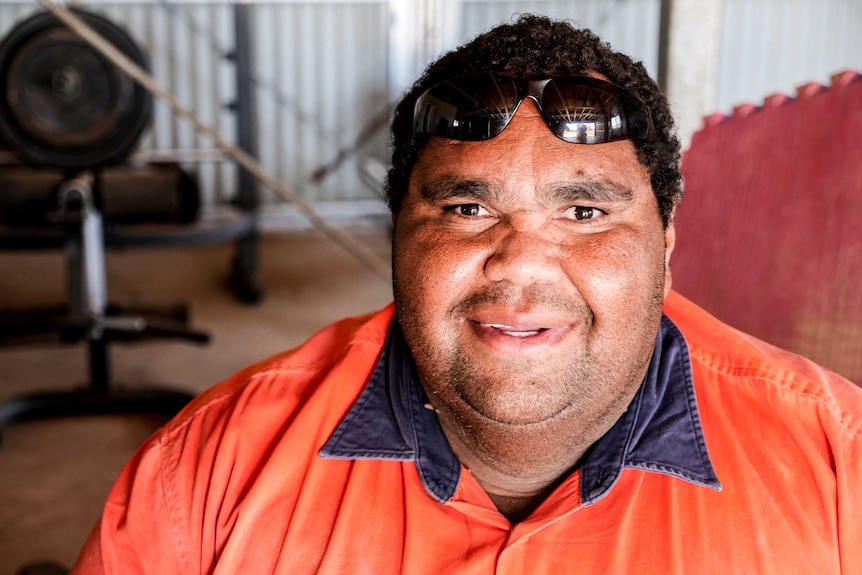 A headshot of an Indigenous man, smiling at the camera. Behind his is gym equipment.