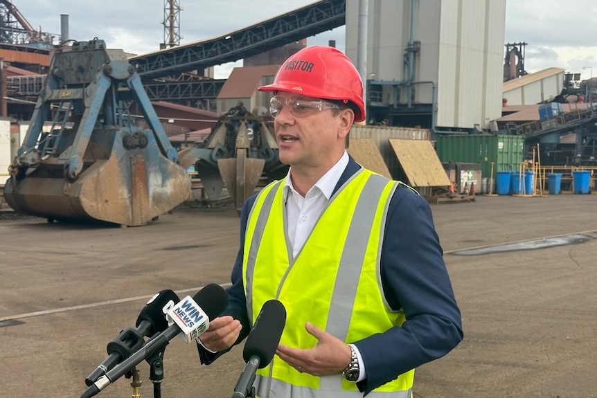 Man in a suit wearing a hi vis jacket and hard hat talking in front of microphones